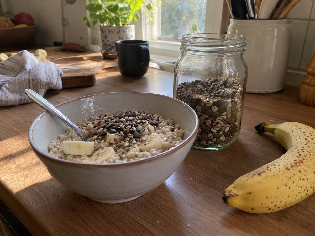 Bowl of porridge oats topped with seeds and banana next to a jar of seeds on a kitchen counter